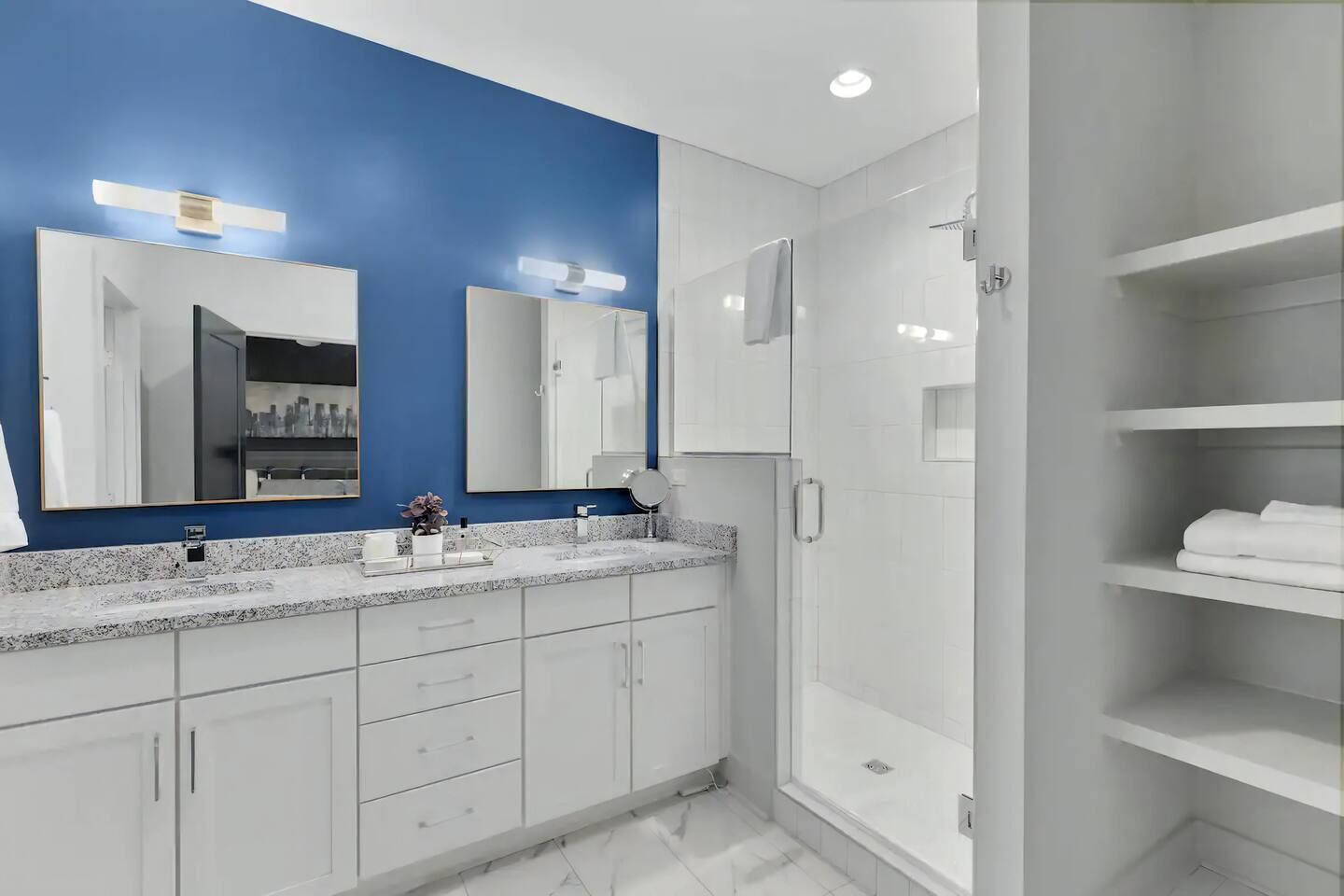 A modern bathroom with a blue accent wall, a large mirror, a walk-in shower, and a built-in vanity with a countertop.