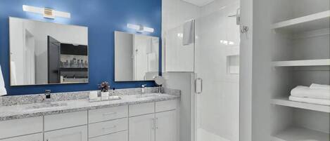 A modern bathroom with a blue accent wall, a large mirror, a walk-in shower, and a built-in vanity with a countertop.