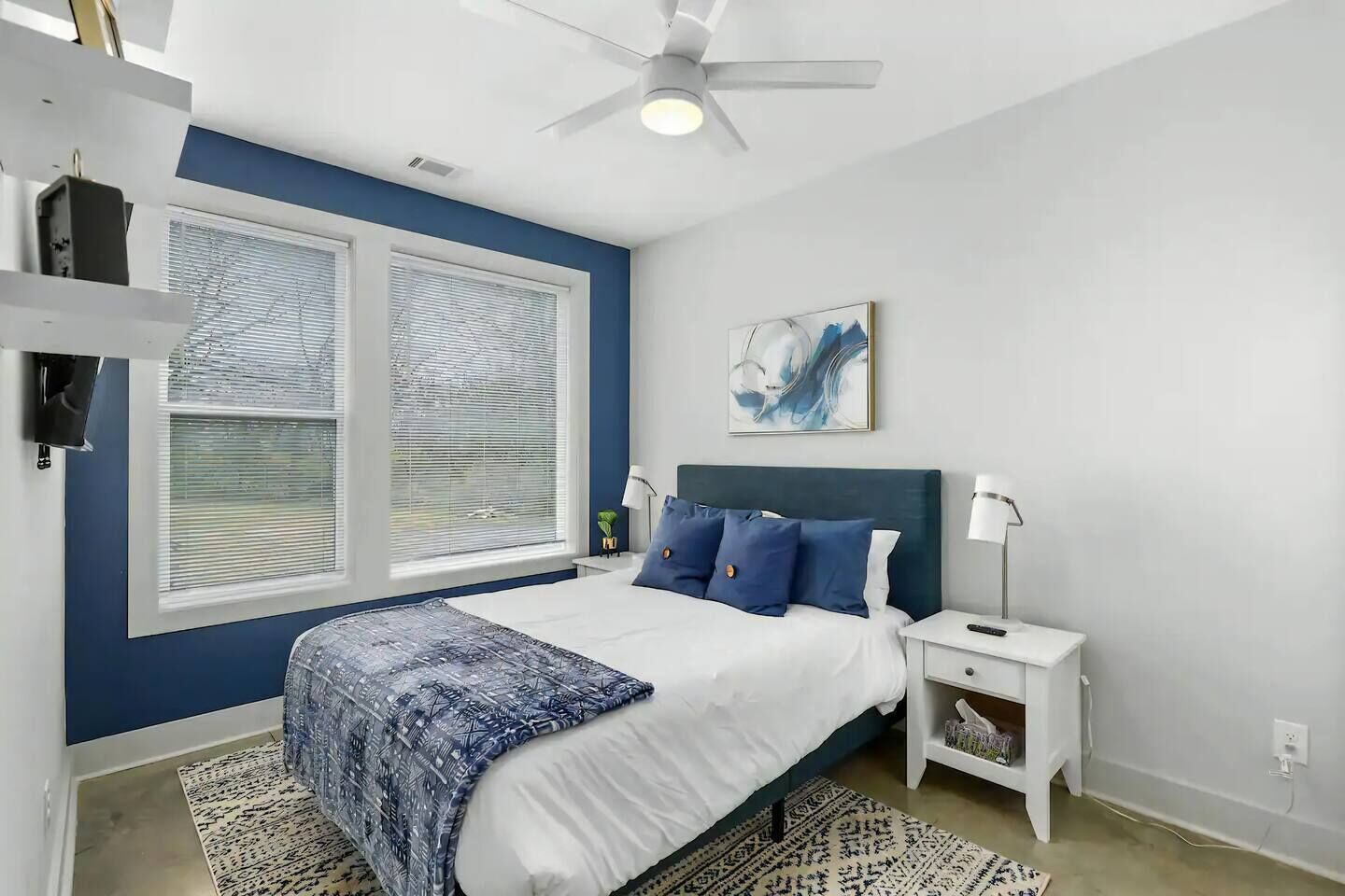 Une chambre à coucher comprenant un lit, des tables de chevet, un ventilateur de plafond, une fenêtre avec des stores et une œuvre d’art accrochée au mur.