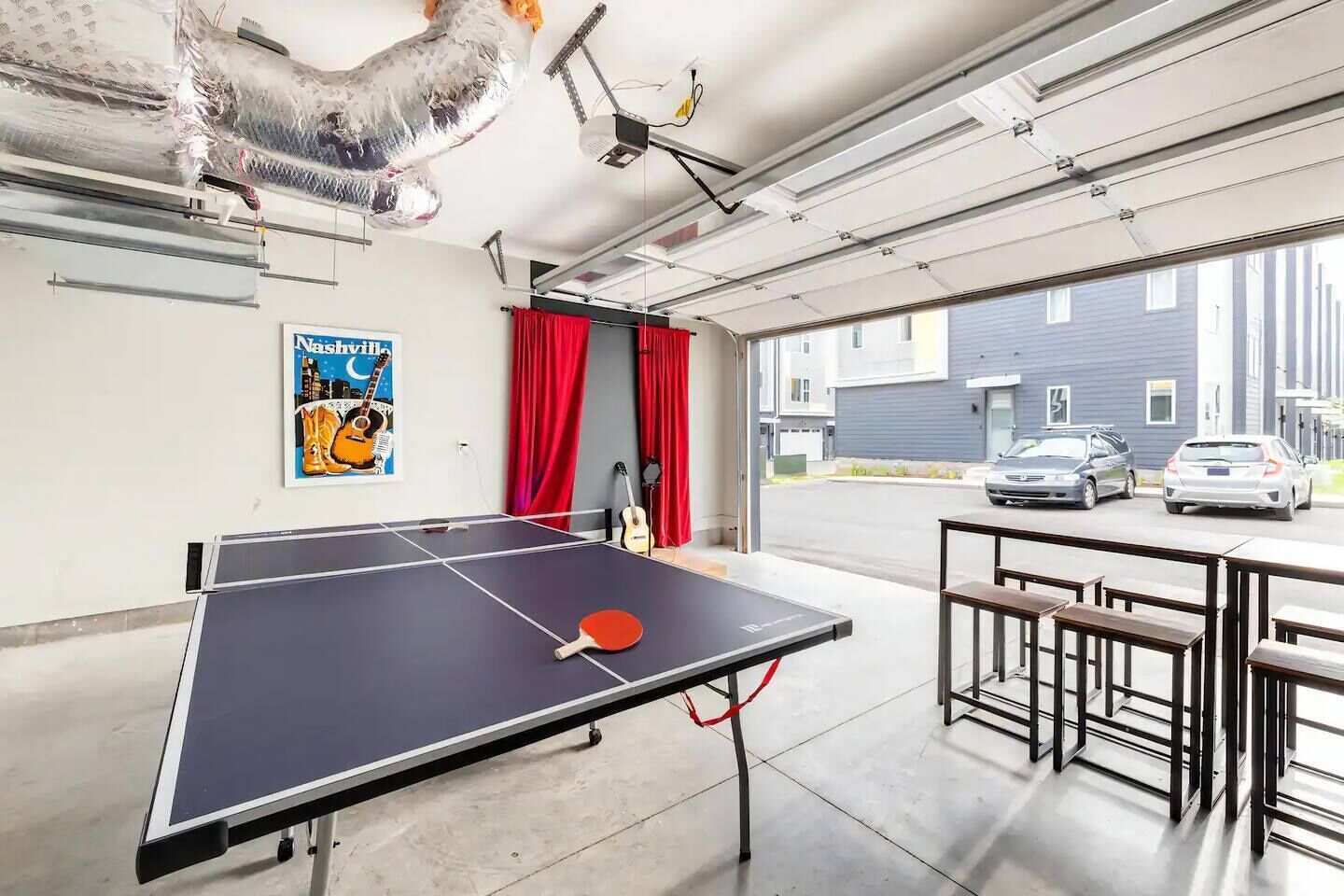 Una mesa de ping pong en una habitación con un ventanal grande, cortinas rojas y una guitarra apoyada en la pared.