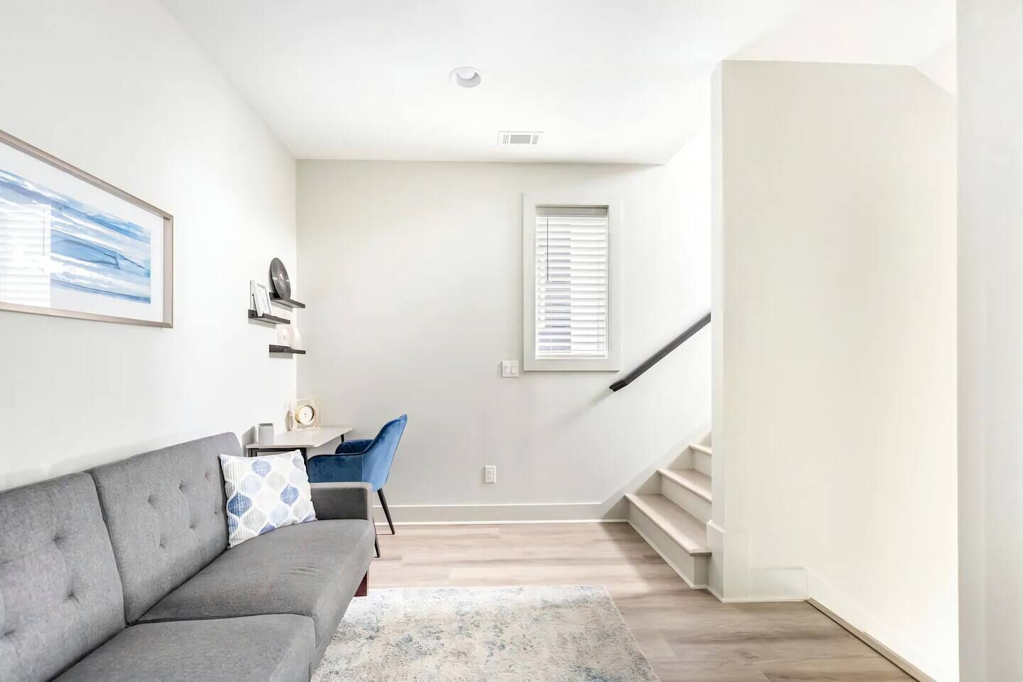 Una sala de estar moderna con un sofá gris, un sillón azul, una mesita y una escalera que lleva a un nivel superior.