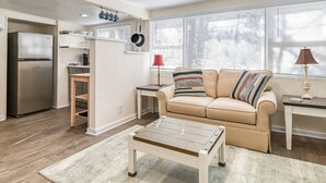A compact living space with a kitchenette, a beige sofa, a coffee table, and a window with blinds. - Little Palm Hideaway Cottage (St. Augustine)