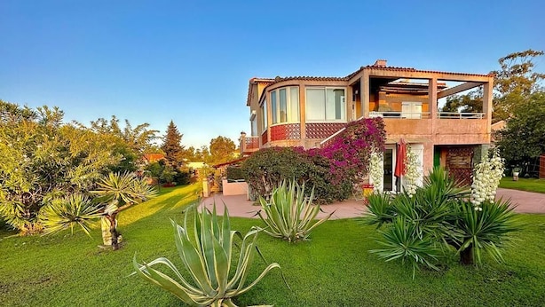 Una casa de dos pisos con balcón, rodeada de céspedes cuidados y diversas plantas.