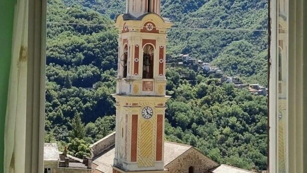 A view through a window with a curtain, featuring a tower with a clock and a cross on top, set against a backdrop of green hills and a clear sky. - La Casa Di Edy (Prato)