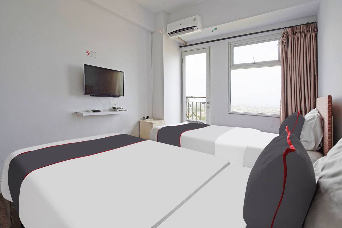 Habitación de hotel con dos camas, televisor, equipo de aire acondicionado, ventana con cortinas y balcón.