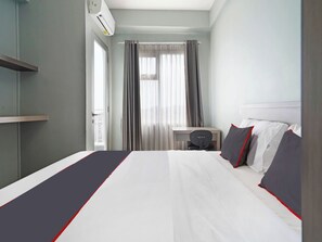 Ein Hotelzimmer mit einem Bett, einem Schreibtisch und einem Stuhl. Es gibt ein Fenster mit Vorhängen, eine Klimaanlage und einen Blick auf Gebäude.