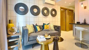 A modern living room with a grey sofa, yellow and blue cushions, a round coffee table, and a kitchen area with a bar counter.