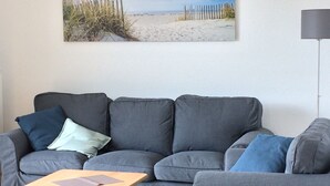 Una sala de estar con un sofá gris, una mesa de centro de madera y un cuadro con motivos playeros en la pared.