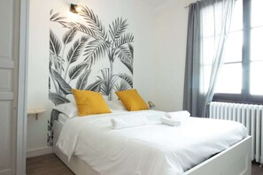 Un lit bien fait, avec du linge de lit blanc et deux coussins jaunes, un mur orné d’un motif de feuilles tropicales et une fenêtre avec des rideaux.