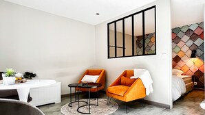 Una habitación de hotel moderna con una cama blanca, un sillón naranja, una pequeña mesa redonda y un ventanal con cortina de estampado geométrico.