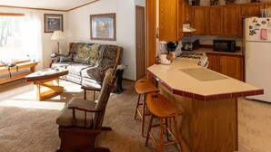 A cozy living room with a sofa, a wooden coffee table, and a rocking chair. The kitchen area includes wooden cabinets, a microwave, and a refrigerator.