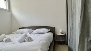 A neatly made bed with white linens and pillows, a bedside table with a lamp, and a window with curtains. - Leana t2 avec parking (Annecy)