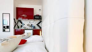 Habitación de hotel compacta con kitchenette, cama con ropa de cama blanca y almohadas rojas, y un espejo en la pared.