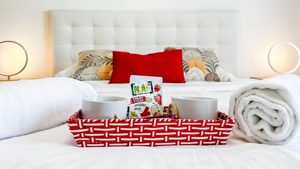 Una cama bien hecha con cabecera blanca, almohadas rojas y una cesta de regalo con fruta.