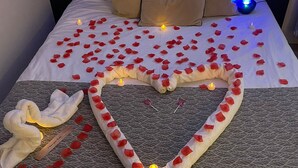 Ein Bett mit Rosenblättern, die in Herzform drapiert sind, ein aufgerolltes Handtuch und eine kleine herzförmige Dekoration auf einer gemusterten Tagesdecke.