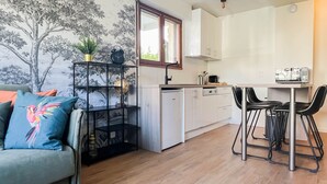 Ein kompakter Wohnbereich mit Küchenzeile, einer Essecke mit zwei Stühlen, einem Sofa mit farbigem Kissen und einem an der Wand befestigten Regal mit Dekoration.
