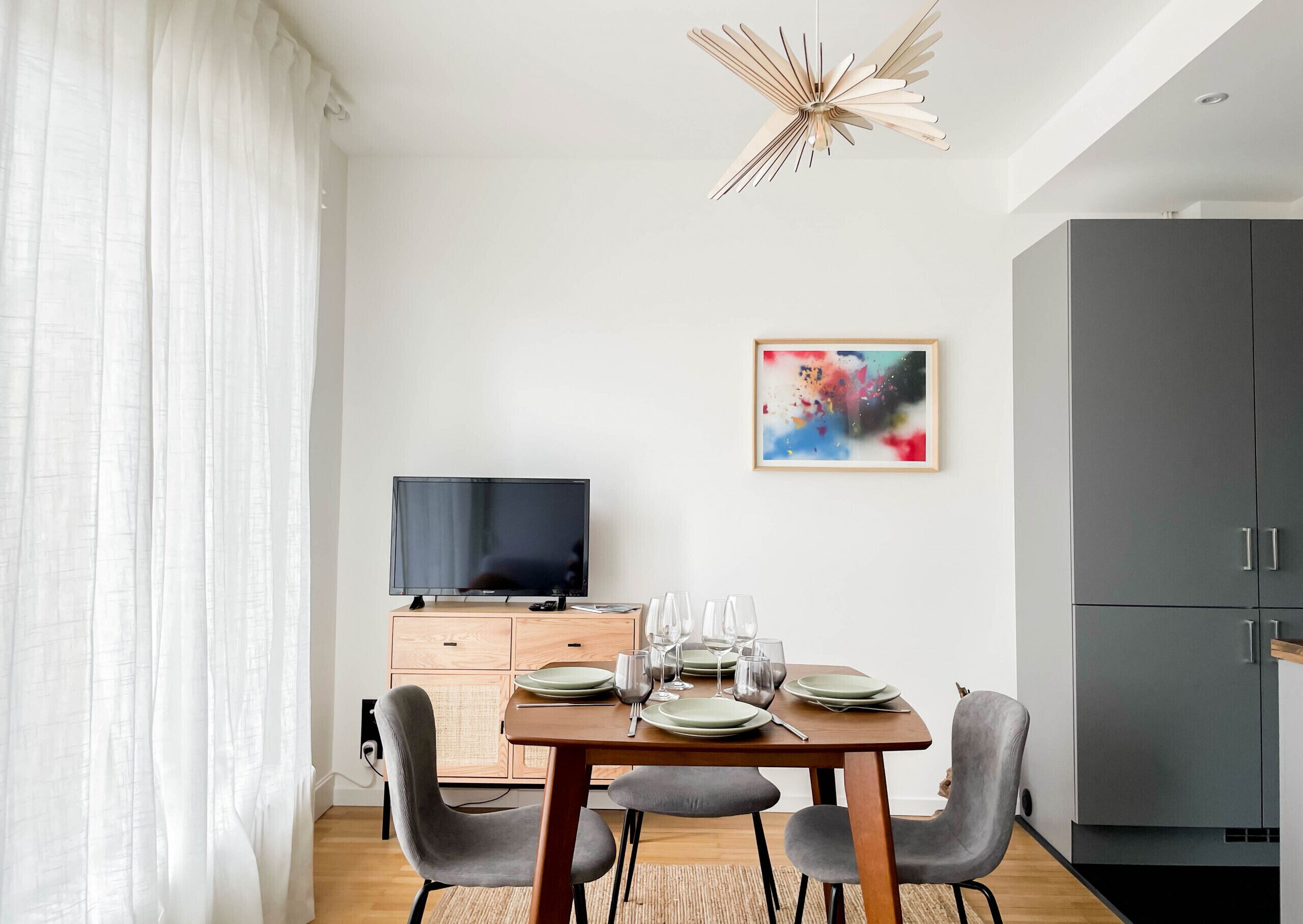 Un espace repas moderne avec une table en bois dressée pour quatre personnes, un téléviseur posé sur un meuble et un tableau abstrait aux couleurs vives accroché au mur.