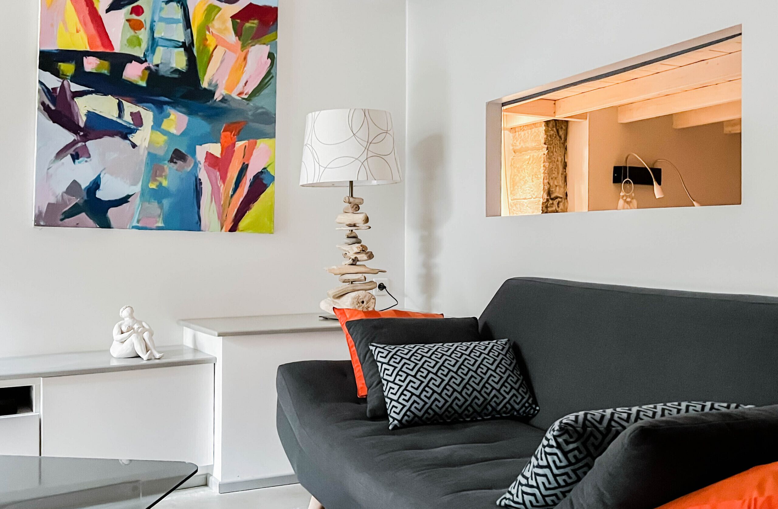 Een moderne woonkamer met een zwarte bank, gekleurde kussens, een witte bijzettafel, een kleurrijk abstract schilderij, een bijzondere lamp, en een ingelijste foto aan de muur.