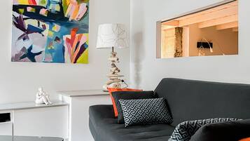 Een moderne woonkamer met een zwarte bank, gekleurde kussens, een witte bijzettafel, een kleurrijk abstract schilderij, een bijzondere lamp, en een ingelijste foto aan de muur.