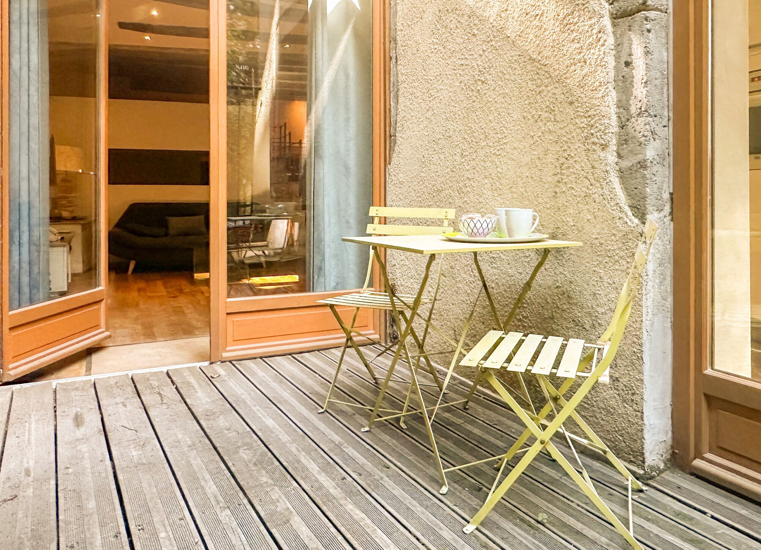 Um deck de madeira com uma pequena mesa e cadeira, uma porta de vidro que dá para o interior, e uma parede com textura.