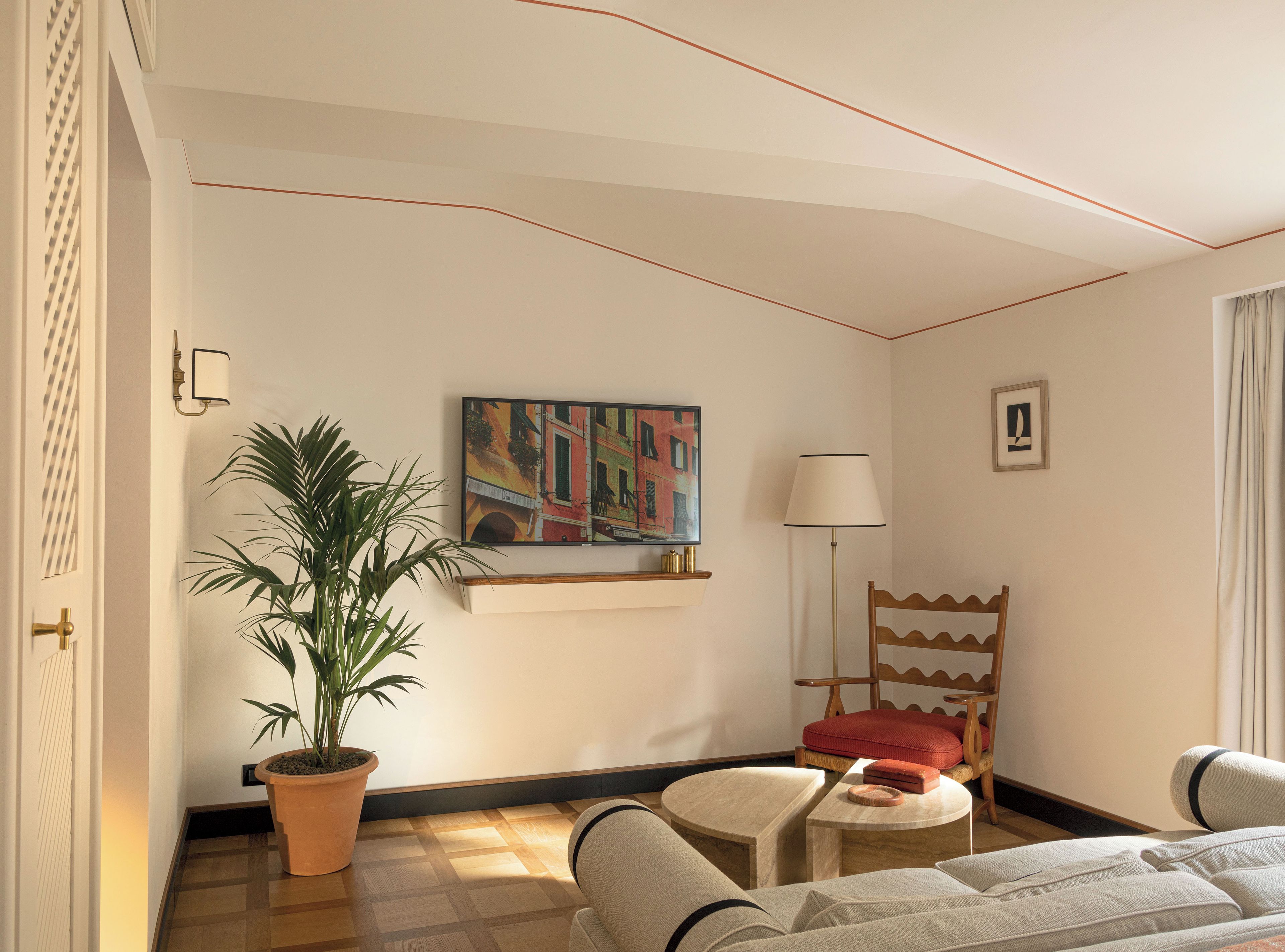 A hotel room with a bed, a chair, a table, a potted plant, a painting, and a lamp.