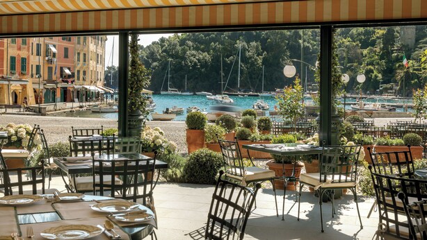Un área de comedor al aire libre con mesas y sillas, con vistas a un puerto deportivo con barcos y un pueblo costero al fondo.