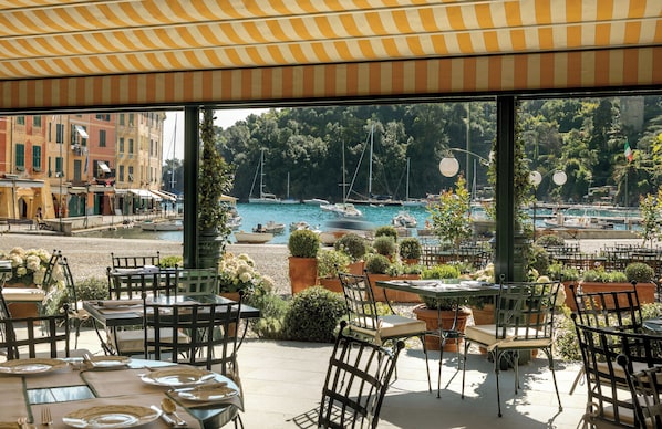 An outdoor dining area with tables and chairs, overlooking a marina with boats and a coastal town in the background.