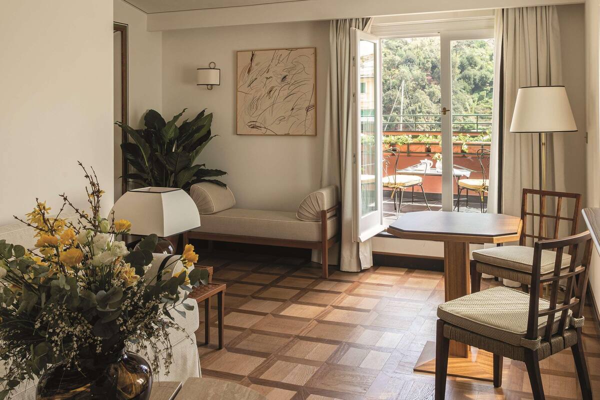 A cozy living room with a sofa, a dining table, and chairs. There is a window with curtains, a plant, and a painting on the wall.