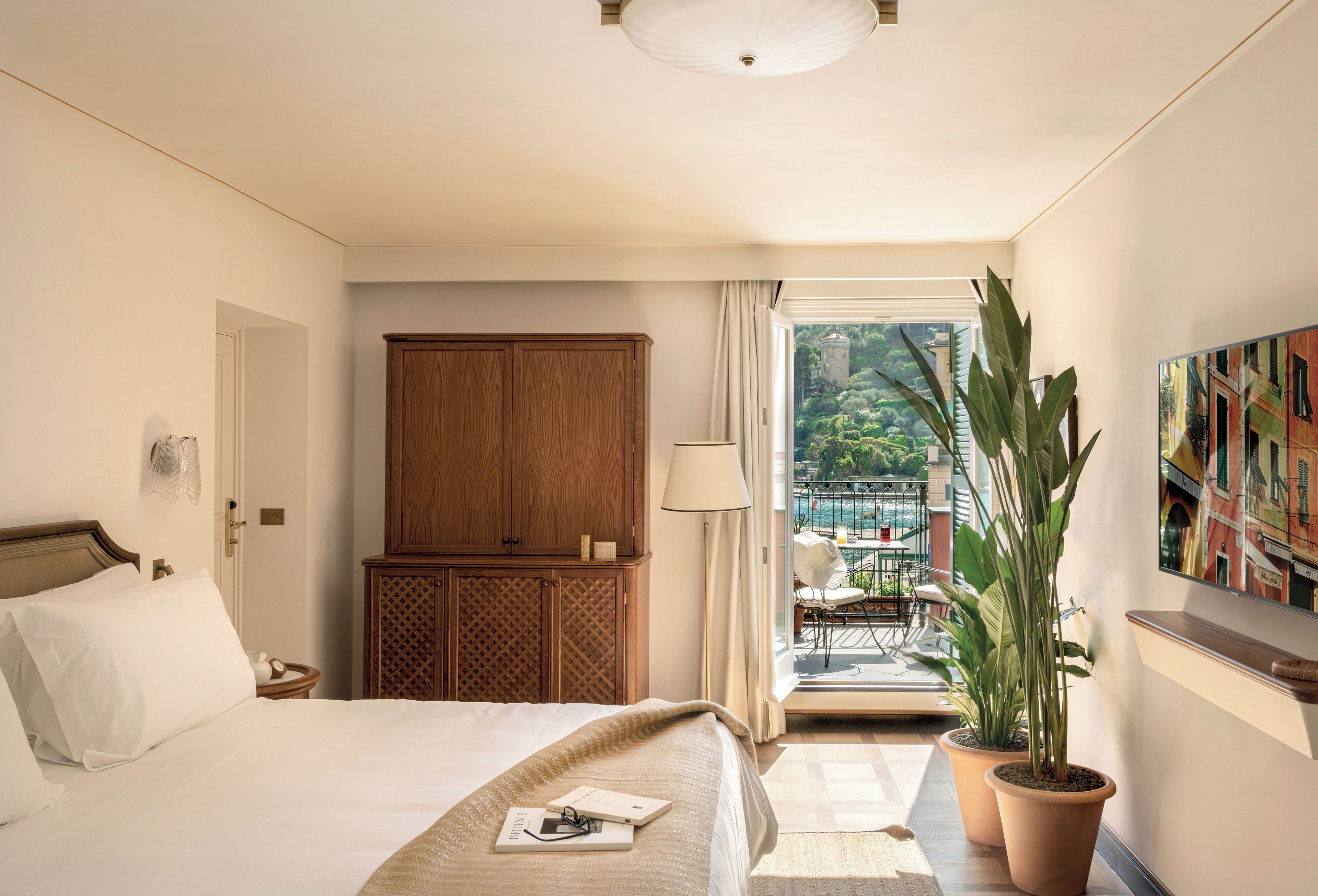 A hotel room with a bed, wooden wardrobe, potted plants, and a view of the outside balcony.