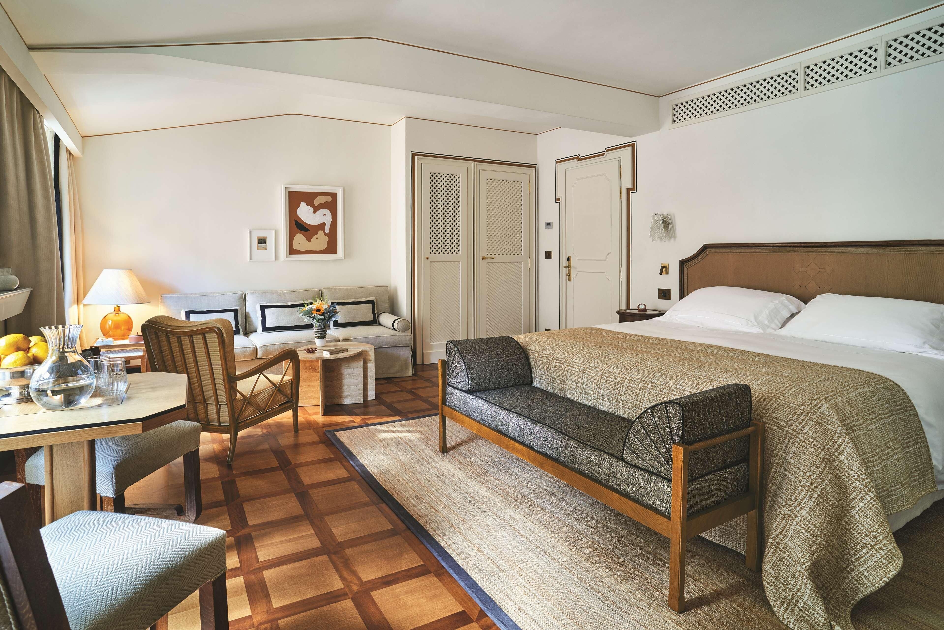 A hotel room with a bed, a sofa, a dining table with chairs, a lamp, and a decorative wall piece.