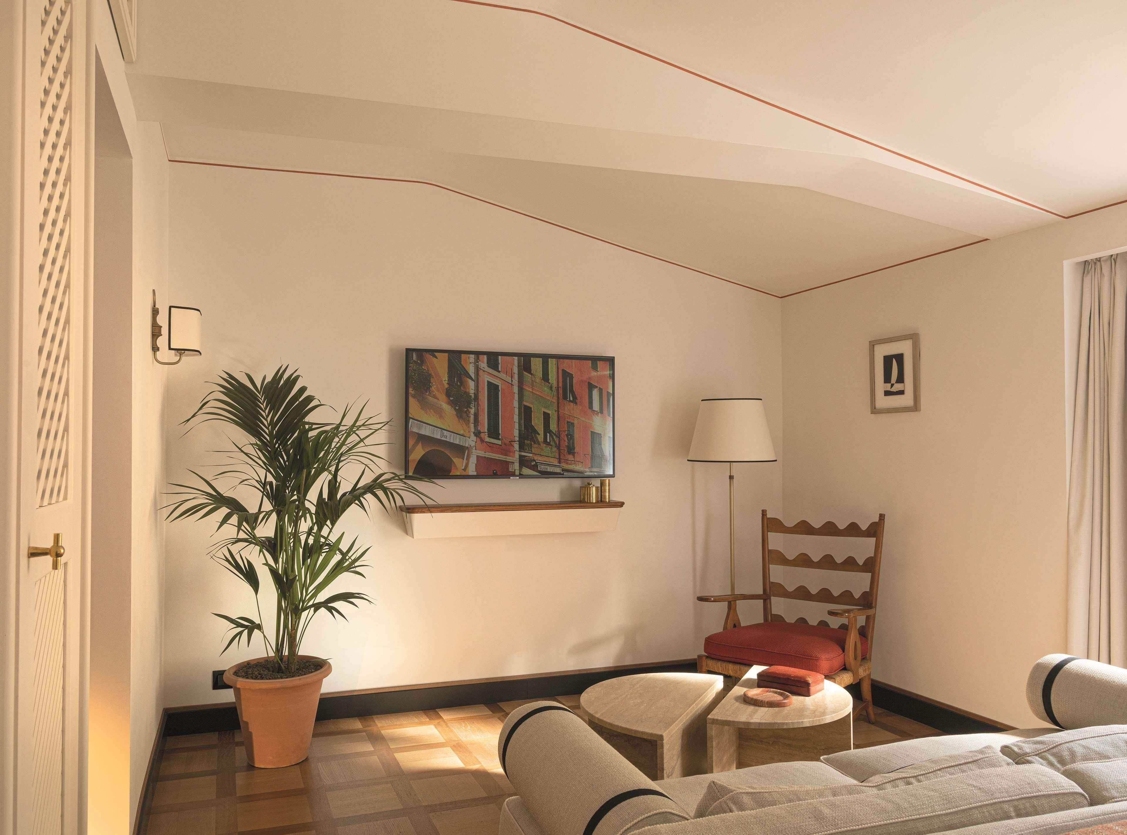 Habitación de hotel con una planta grande en maceta, un cuadro llamativo, una lámpara, una silla con cojín rojo y una cama con cabecera blanca.