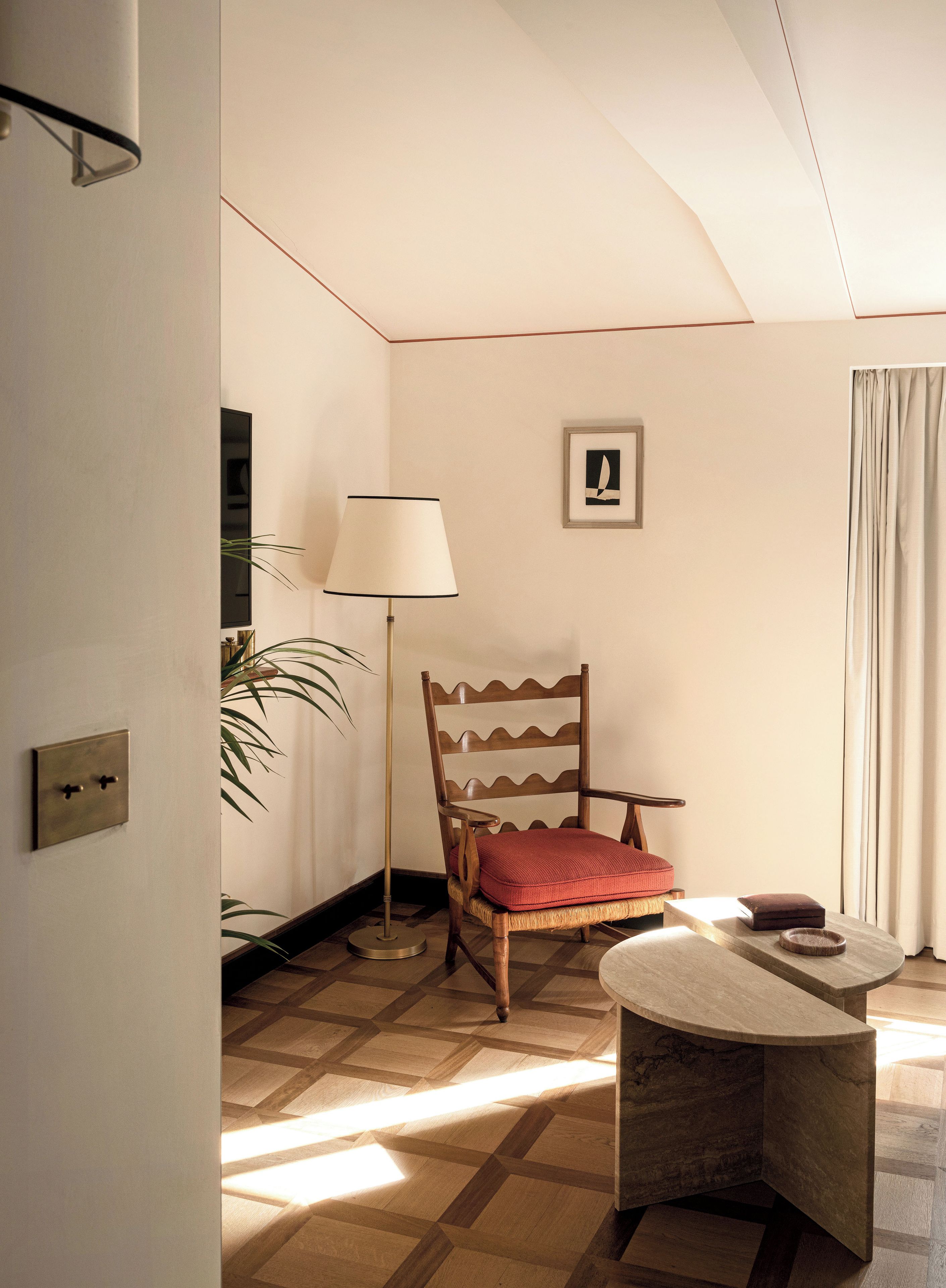 Een kamer met een houten stoel, een tafel, een vloerlamp en een schilderij aan de muur.