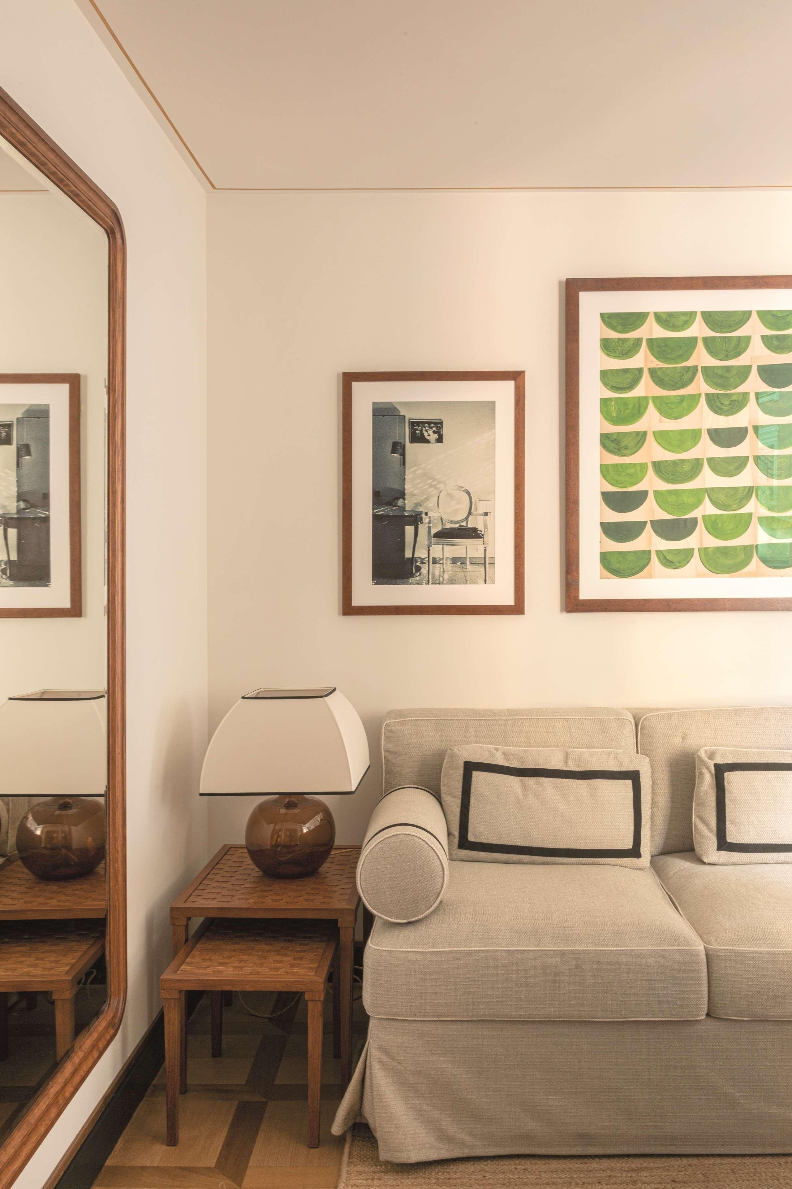 Una sala de estar con un sofá color beige, una pequeña mesa de madera con una lámpara y cuadros en la pared.
