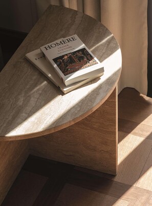 A book titled "HOMÈRE" on a curved wooden table with a marble top, positioned near a window with sunlight casting shadows. - Splendido Mare, A Belmond Hotel, Portofino (Santa Margherita Ligure)