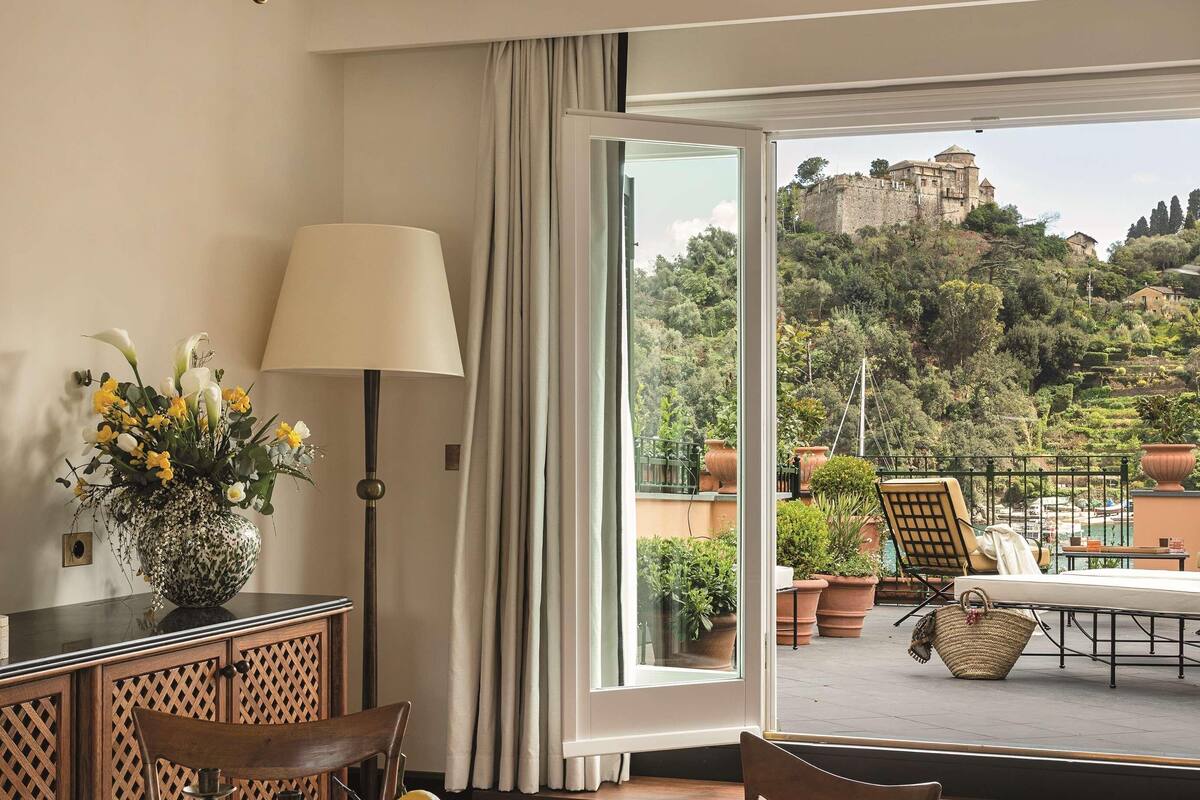A room with a round table, chairs, a vase with flowers, a lamp, and a view of a castle through a large window.