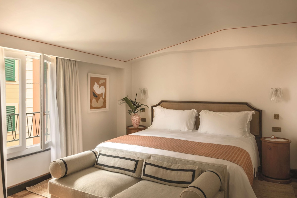 A neatly arranged bedroom with a large bed, a bedside table with a lamp, a potted plant, and a window with curtains.