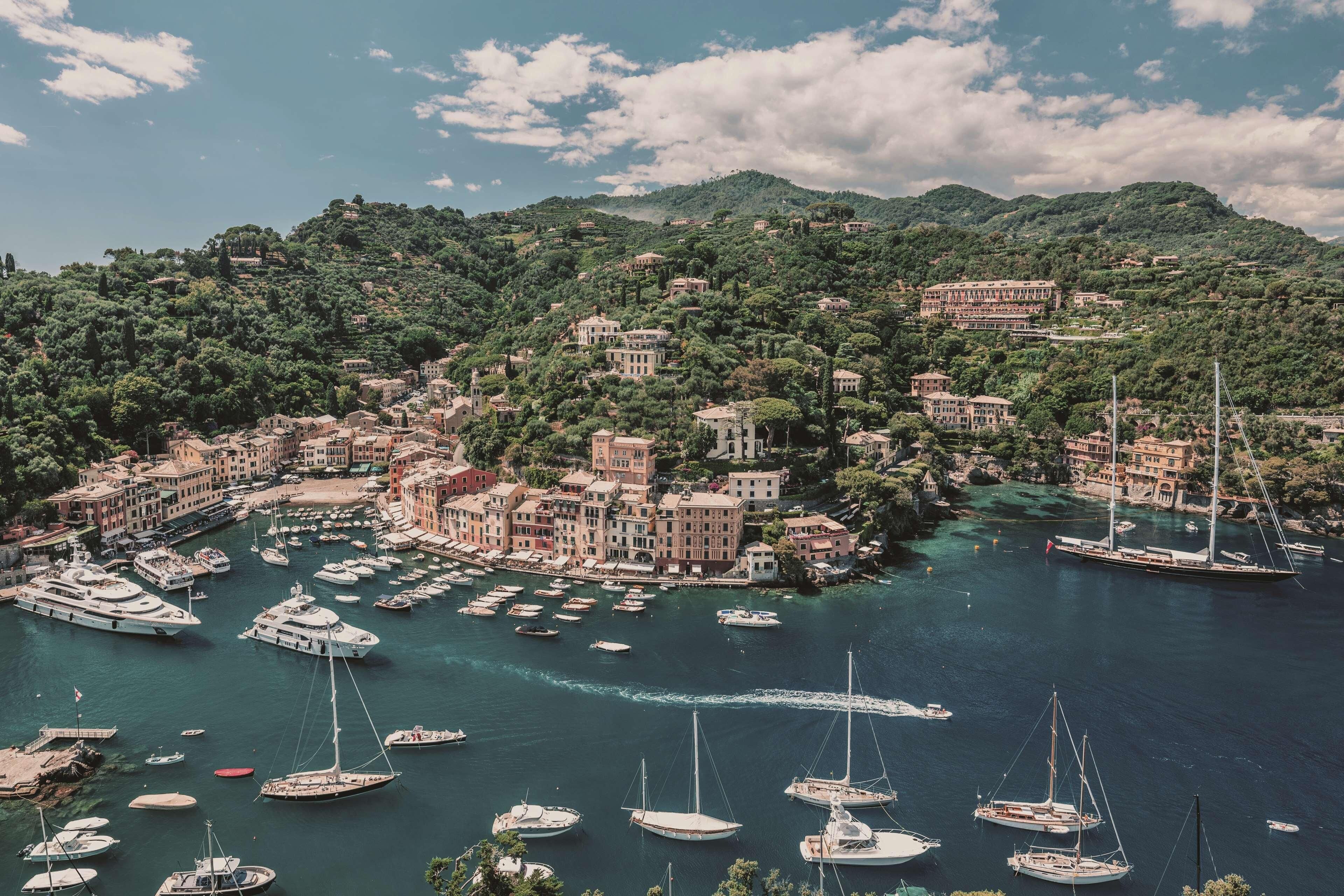 A coastal town with numerous boats docked in a marina, surrounded by lush green hills and historic buildings.