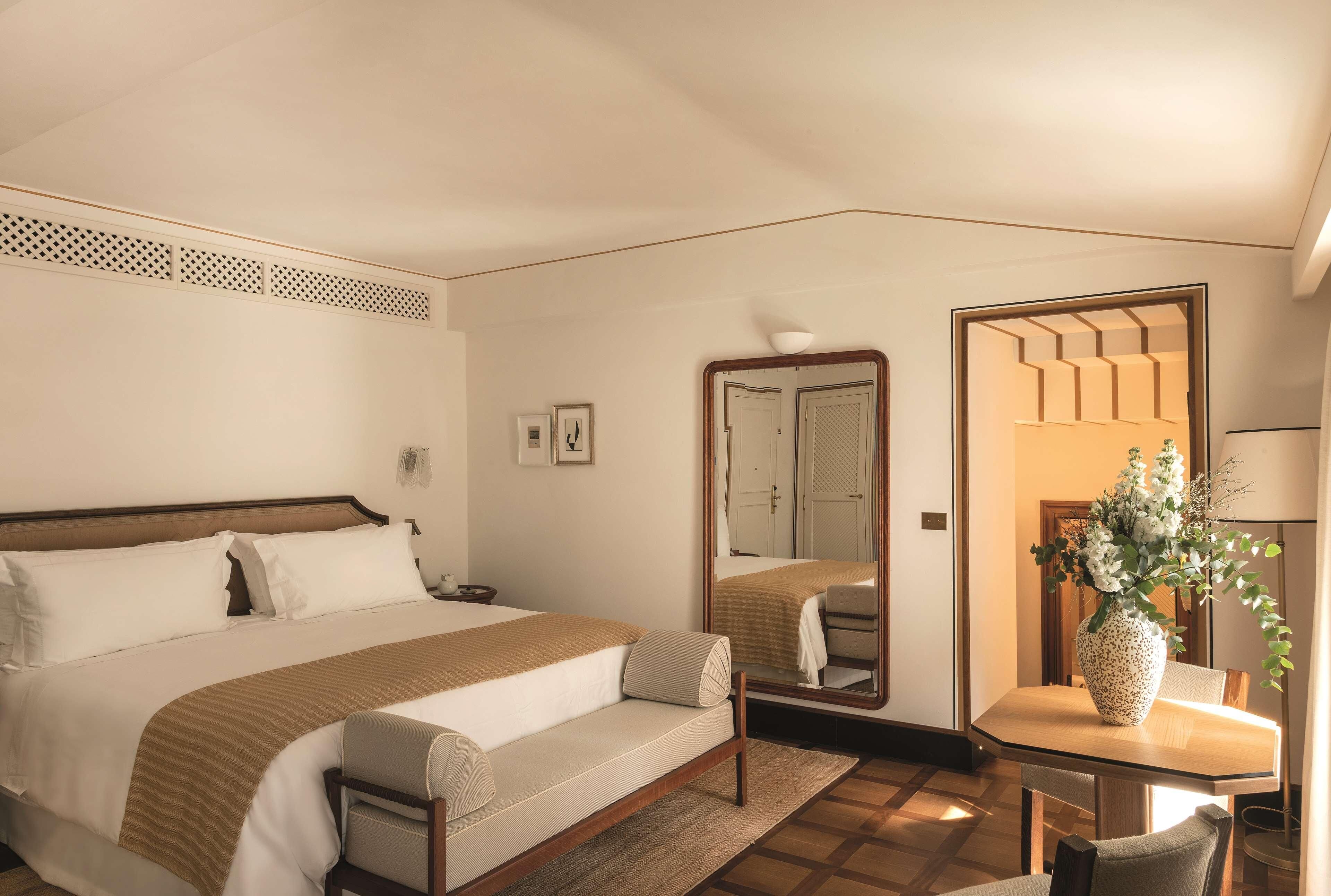 Een hotelkamer met een bed, nachtkastje, spiegel, stoel en een tafeltje met een bloemenvaas.