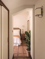 Corridoio di un albergo con un letto, un comodino, una pianta in vaso e un punto luce a parete.