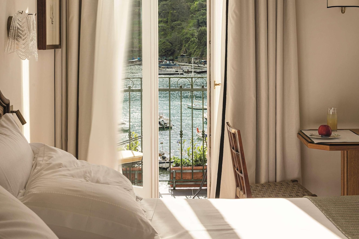 A hotel room with a bed, a table with a red object, and a view of a lake and trees through a window.