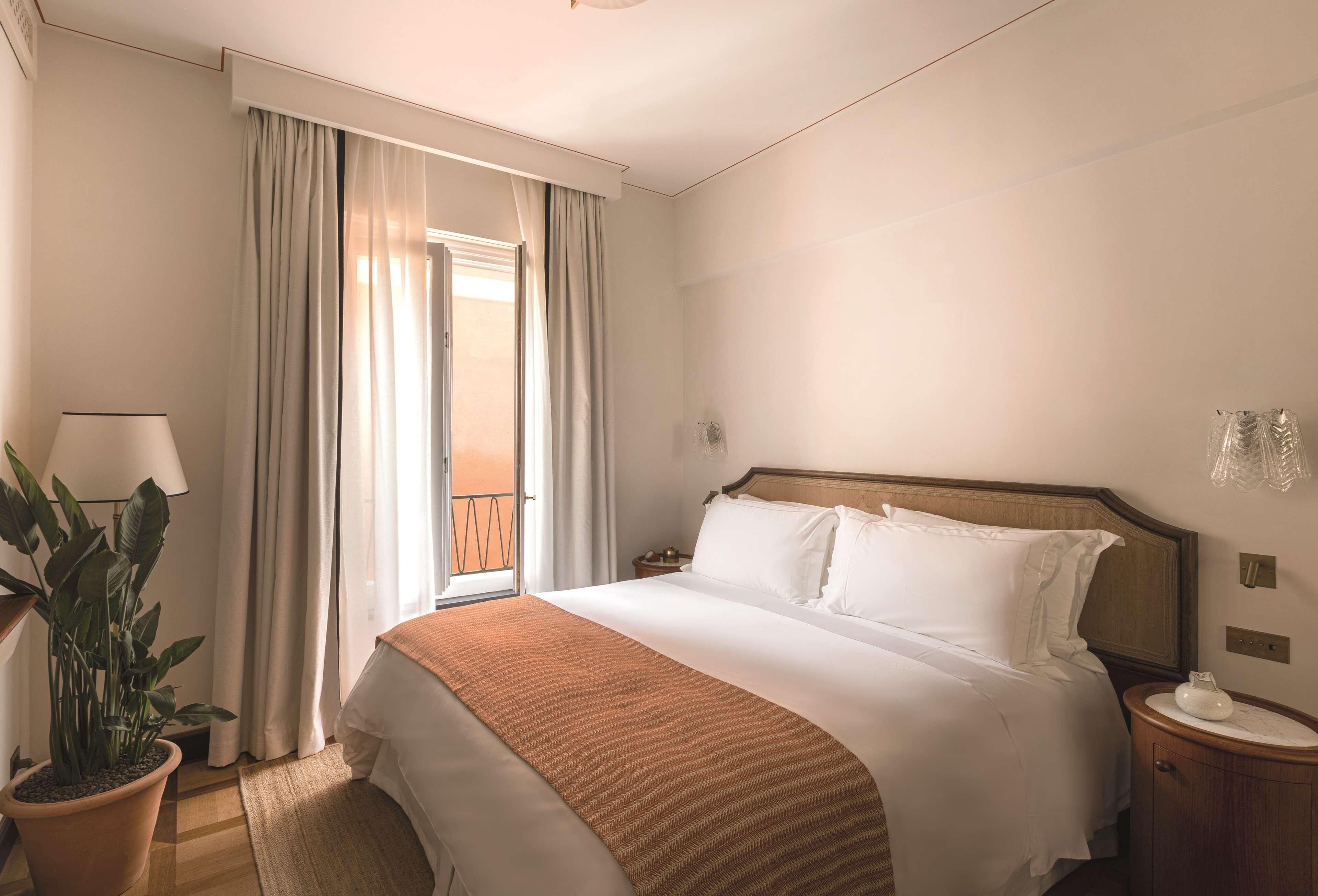 A hotel room with a bed, a nightstand, a lamp, a potted plant, and a view of the outdoors.