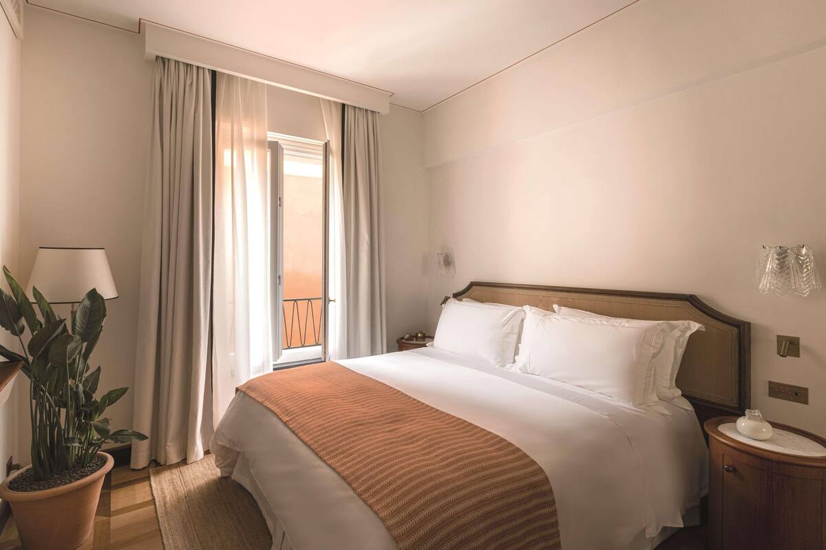 A hotel room with a bed, a nightstand, a lamp, a potted plant, and a view of the outdoors.