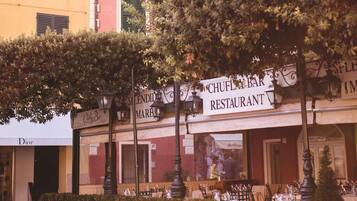 A restaurant with outdoor seating, a building with a red facade, and a tree in the foreground.