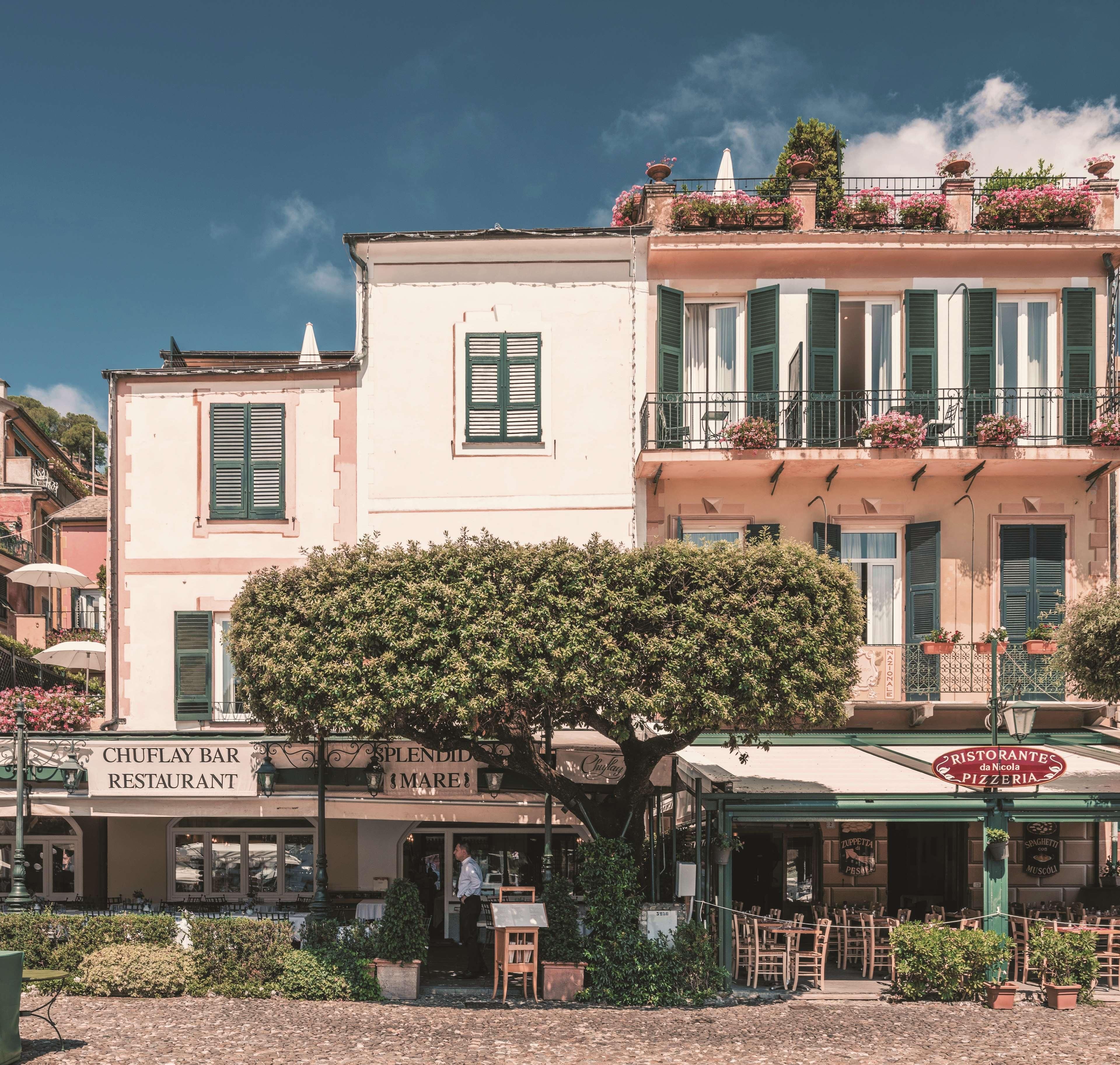 Uma fileira de edifícios com um restaurante chamado "CHUFLAY BAR RESTAURANT" e uma pizzaria "RISTORANTE da Nicola PIZZERIA", ambos com mesas ao ar livre.