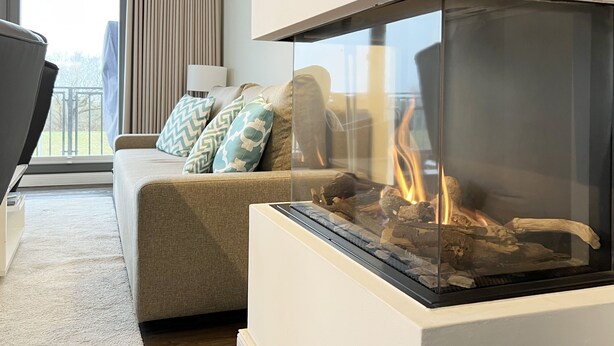 Ein modernes Wohnzimmer mit Kamin aus Glas, einem beigefarbenen Sofa mit gemusterten Kissen und Blick ins Freie durch große Fenster.
