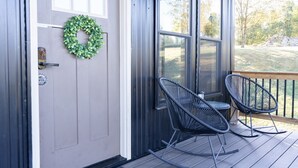 A porch with two rocking chairs, a door with a wreath, and a view of the outdoors.