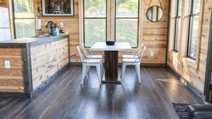 A wooden cabin interior with a dining table and chairs, large windows, and a kitchen area.