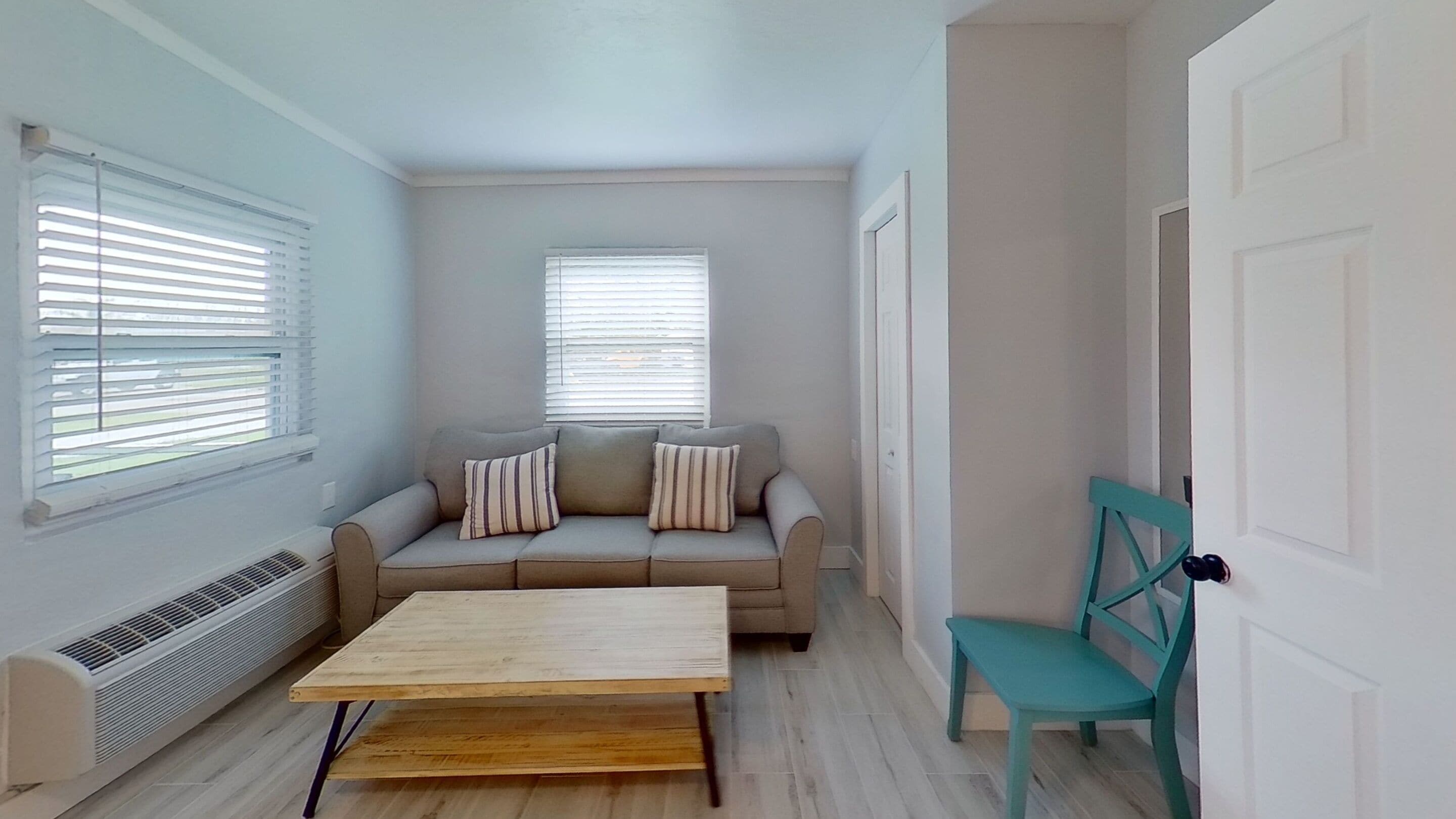 Una habitación pequeña y bien iluminada con un sofá, una mesa de madera y una silla color turquesa.