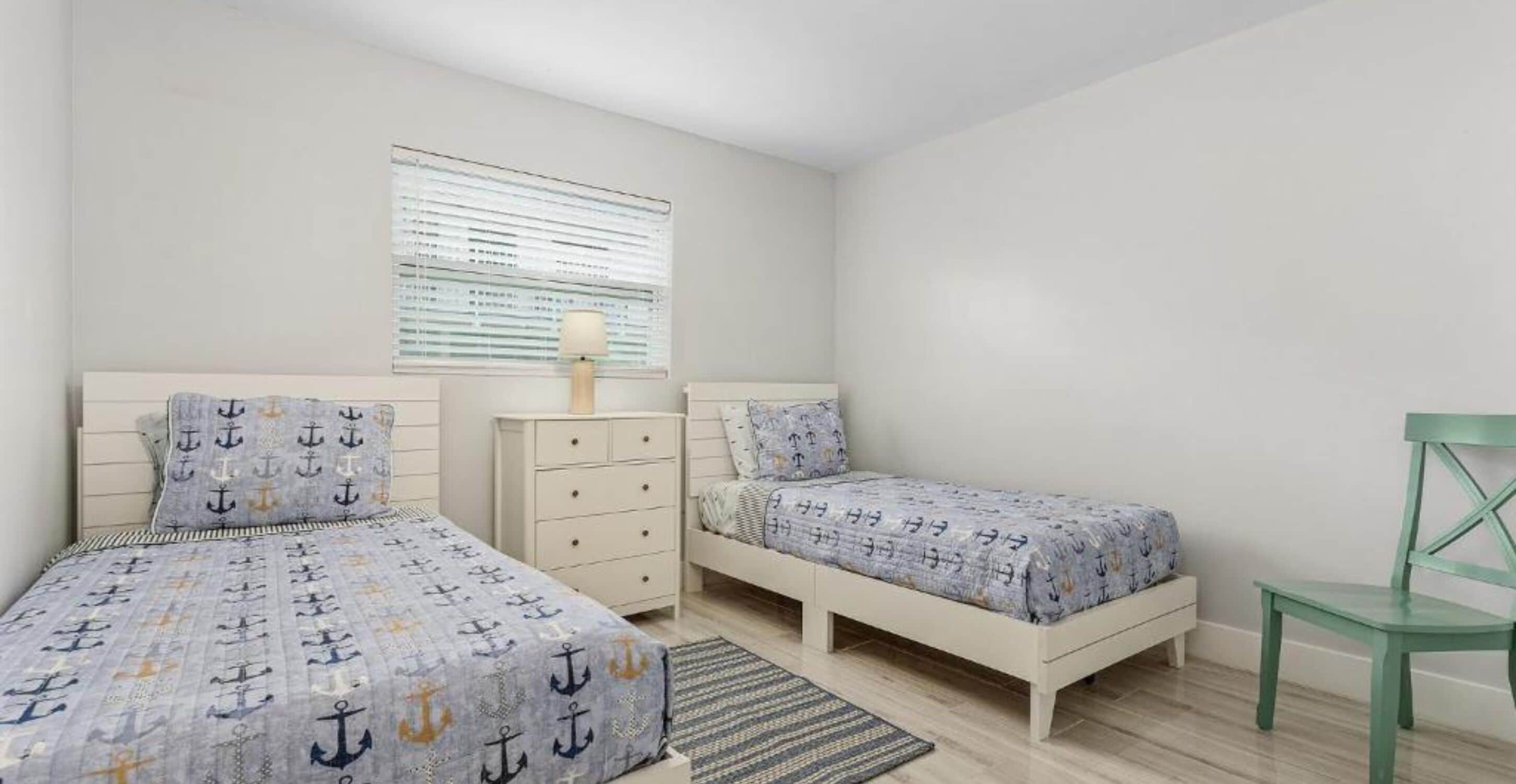 Une chambre avec deux lits, une commode, une chaise et une fenêtre avec des stores.