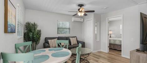 Un salon moderne avec une table ronde en verre, des fauteuils turquoise, un canapé gris avec des coussins à rayures et un ventilateur de plafond.